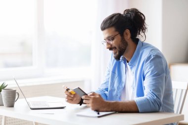 Genç Arap adam evde akıllı telefon ve kredi kartı kullanıyor, internetten alışveriş yapıyor.