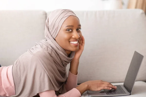 Retrato de jovem negra no hijab deitado no sofá e usando laptop em casa, tendo trabalho freelance, espaço de cópia — Fotografia de Stock