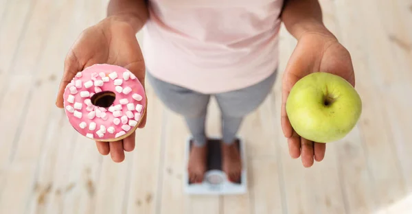 Gezond versus ongezond voedsel. Zwarte vrouw met appel en donut op schubben, met keuze van haar maaltijd, boven het zicht — Stockfoto