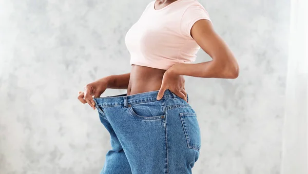 Oigenkännlig svart kvinna i överdimensionerade jeans visar resultat av hennes viktminskning kost eller fettsugning, ljus bakgrund — Stockfoto