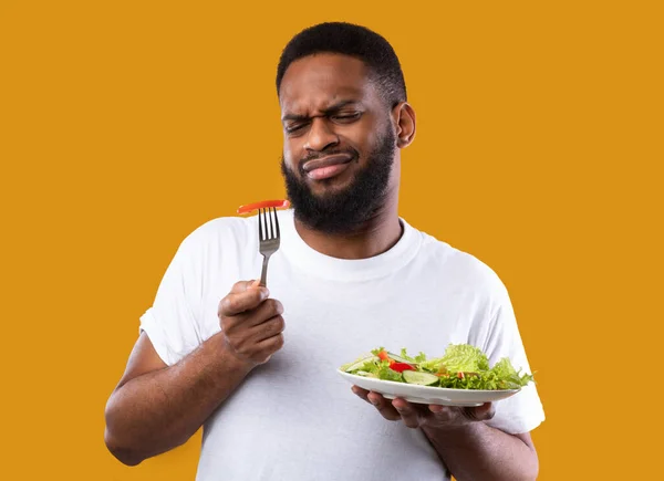 Homem afro-americano infeliz comendo salada de legumes sobre fundo amarelo — Fotografia de Stock