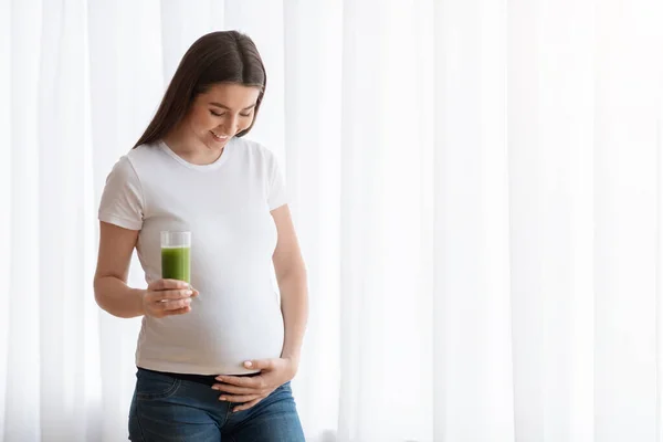 Щаслива молода вагітна жінка п'є зелений овочевий сік або смузі вдома — стокове фото