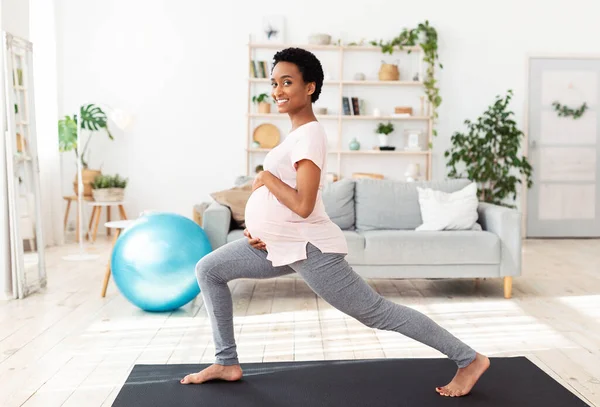 Embarazo activo y deportivo. Mujer negra embarazada alegre haciendo embestidas, estirando las piernas, haciendo ejercicio en casa —  Fotos de Stock