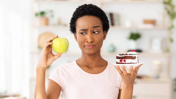 ケーキの代わりに果物を食べたくないという疑いでアップルを見ている若い黒人女性 — ストック写真