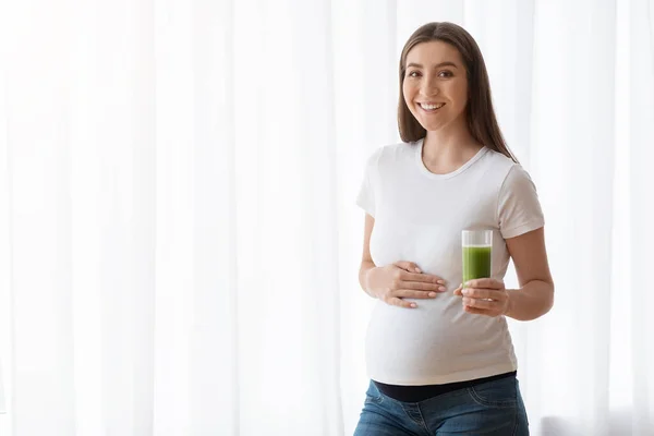 Усміхнена вагітна жінка зі склом зеленого овочевого смугастого стоїть біля вікна — стокове фото