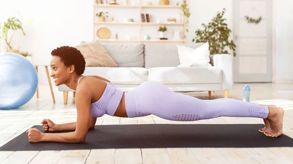 Vista lateral de la señora negra haciendo tabla de codo en estera de yoga en la sala de estar —  Fotos de Stock