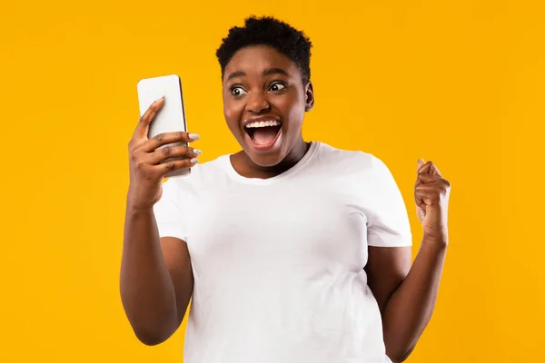 Mulher Africana segurando Smartphone balançando punhos em alegria, fundo amarelo — Fotografia de Stock