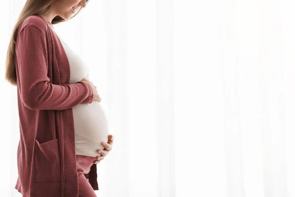 Concepto de embarazo. Feliz joven embarazada abrazando el vientre mientras está de pie cerca de la ventana — Foto de Stock