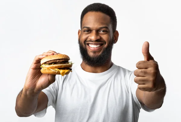 Preto homem segurando hambúrguer e gestos polegares-up sobre fundo amarelo — Fotografia de Stock