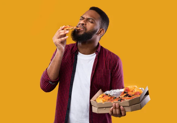 Africano americano homem desfrutando de comer pizza em pé sobre fundo amarelo — Fotografia de Stock