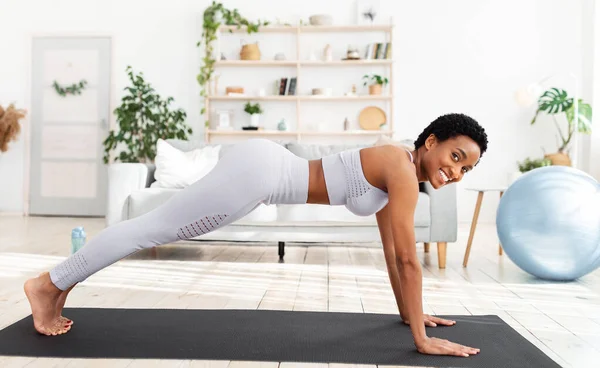 Bastante joven negra haciendo entrenamiento doméstico en esterilla de yoga, de pie en tabla, fortaleciendo los músculos abdominales en casa — Foto de Stock