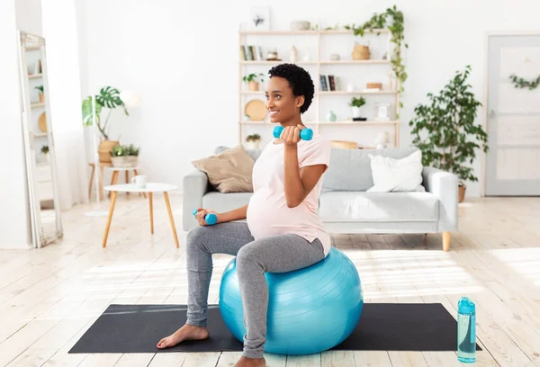 Mujer negra embarazada sentada en una pelota de fitness, haciendo ejercicio con pesas en su gimnasio en casa — Foto de Stock