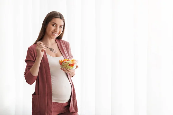 Nutrição durante a gravidez. Sorrindo senhora grávida comendo salada de legumes frescos perto da janela — Fotografia de Stock