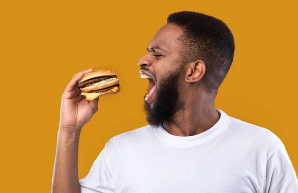 Cara Africano com fome mordendo hambúrguer posando em fundo amarelo, Vista lateral — Fotografia de Stock