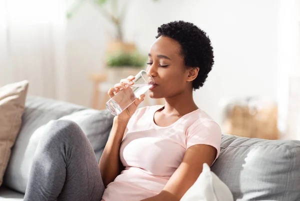 Hidratación para un buen concepto de salud. Bastante joven dama negra teniendo un vaso de agua cristalina en el sofá en casa —  Fotos de Stock