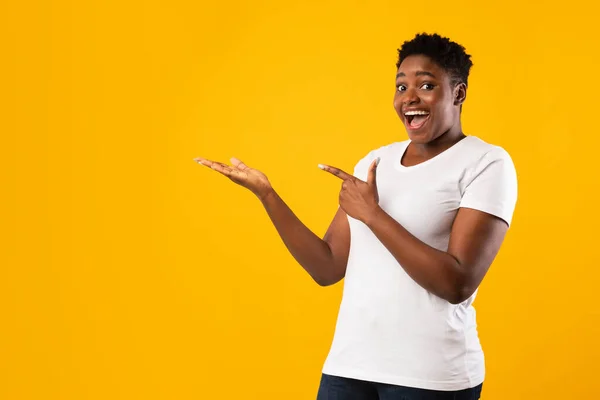 Joyful Plus-Size Africano senhora apontando dedo segurando algo, fundo amarelo — Fotografia de Stock