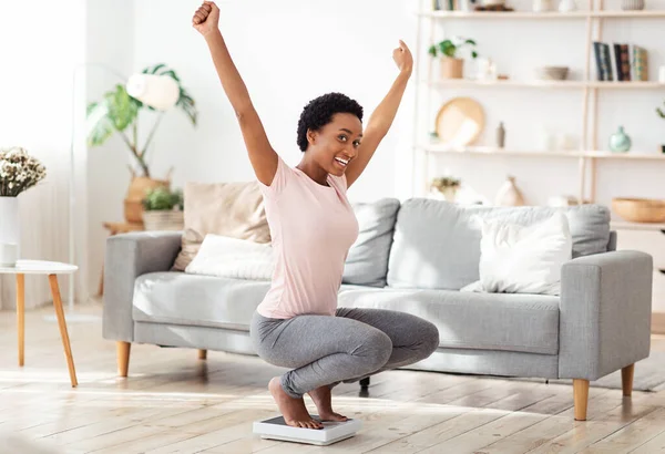 Молодая черная женщина сидит на весах, поднимая руки в волнении, довольная результатом своей диеты потери веса дома — стоковое фото