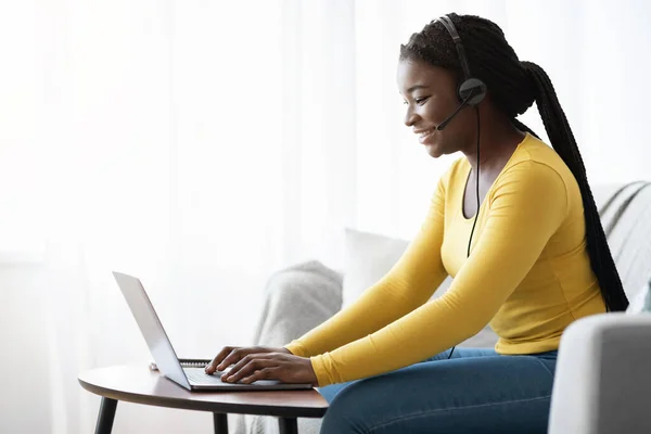 Sorrindo senhora preta com laptop e fone de ouvido trabalhando remotamente em casa — Fotografia de Stock