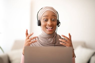 Uzaktan çalışma, iletişim, eğitim. Başörtülü mutlu Müslüman kadın kulaklık takıyor, bilgisayarında video var.