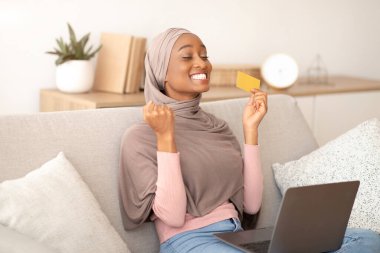 Tesettürlü neşeli siyahi kadın EVET işareti yapıyor, kredi kartı tutuyor, evdeki dizüstü bilgisayarından online alışveriş yapıyor.