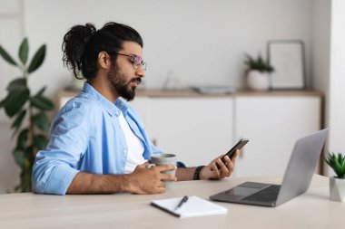 Genç Arap Erkek Serbest Çalışan Akıllı Telefon Kullanıyor ve Kahve İçiyor, İş yerinde Rahatlıyor