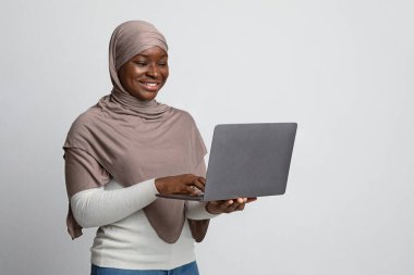 Sırıtkan Müslüman kadın tesettürde dizüstü bilgisayar tutuyor, klavyede daktilo kullanıyor