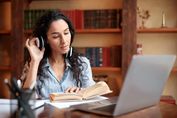 Dame sitzt am Schreibtisch, trägt Kopfhörer, benutzt Computer, liest Buch — Stockfoto