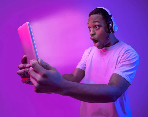 Online Gaming. Black guy in headphones looking at digital tablet with excitement