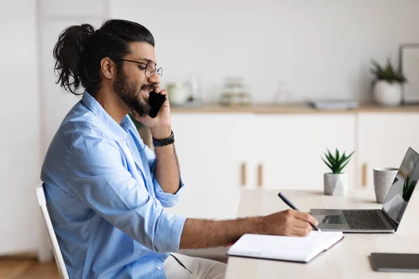 Wesoły Arab Freelancer Guy mówiący przez telefon komórkowy i pisanie notatek do notatnika — Zdjęcie stockowe