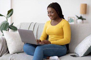 Gülümseyen Afrikalı Amerikalı kadın evdeki koltukta dizüstü bilgisayar kullanıyor.