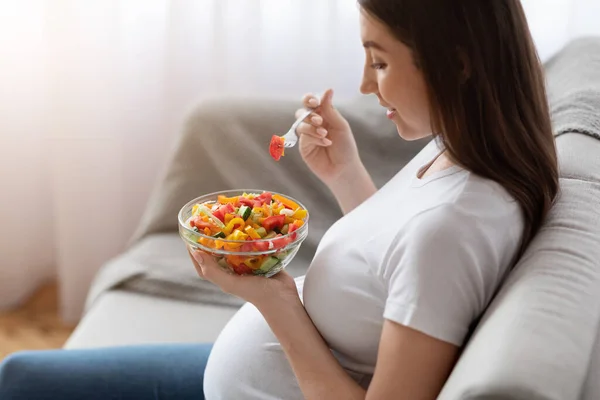 Młoda kobieta w ciąży je świeżą sałatkę warzywną na lunch, siedzi na kanapie — Zdjęcie stockowe