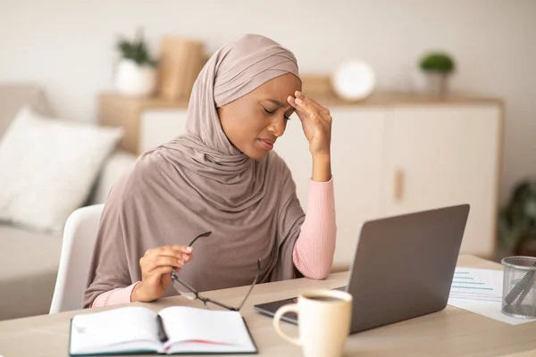 Tensión límite. Mujer negra agotada en hijab cansado después de trabajar en el ordenador portátil durante demasiado tiempo, tener dolor de cabeza en casa — Foto de Stock