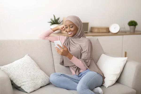 Millenniumi muszlim hölgy hidzsábon mobiltelefonnal otthon, üzenetküldés vagy böngészés a közösségi média, ül a kanapén beltérben — Stock Fotó