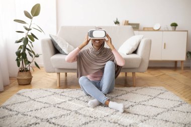 Oturma odasında video oyunu oynayan tesettürlü pozitif siyahi kadın, VR gözlük kullanıyor.