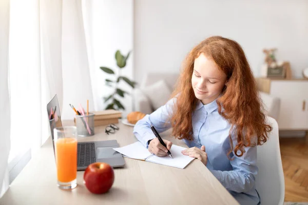 Girl sitting at table, using laptop, writing in her notebook — Stock Photo, Image