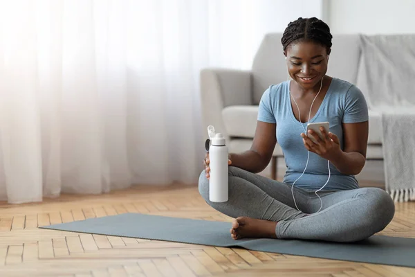 Черная леди расслабляется со смартфоном на коврике для йоги после тренировки дома — стоковое фото