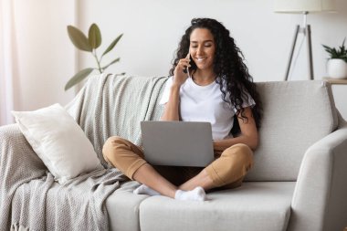 Evde çalışan, dizüstü bilgisayar ve akıllı telefon kullanan genç bir bayan.