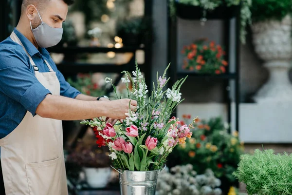 Negócios de flores e criação de buquê. Millennial proprietário masculino em avental e máscara protetora faz arranjo elegante — Fotografia de Stock