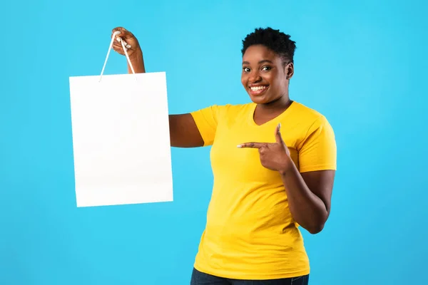 Mujer negra feliz mostrando la bolsa de compras de pie sobre fondo azul —  Fotos de Stock