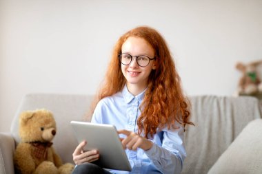 Gözlüklü kız dijital tablet kullanıyor, kanepede oturuyor.