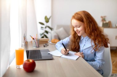 Masada oturan, dizüstü bilgisayar kullanan, deftere yazan bir kız.