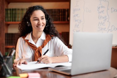 Öğrenci masada oturuyor, dizüstü bilgisayar kullanıyor ve deftere yazıyor.