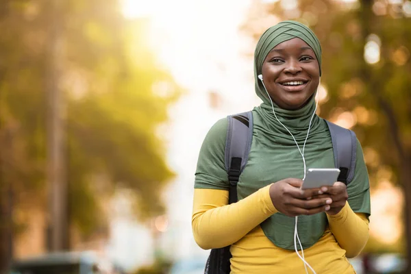 Cheerful Black Muslim Lady In Hijab Walking Outdoors Listening Music On Smartphone