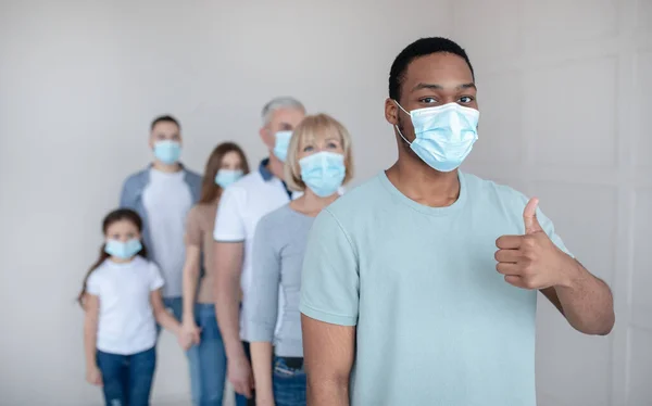 Black guy in face mask waiting in queue for coronavirus vaccination à la clinique, montrant pouce vers le haut geste, copier l'espace — Photo