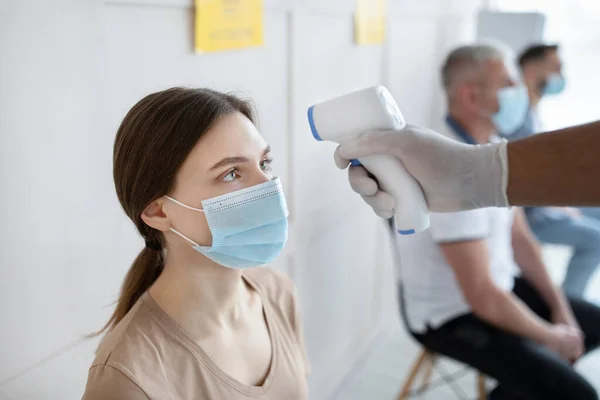 Jovem mulher em máscara facial esperando por imunização covid-19 no hospital, médico verificando sua temperatura com termômetro — Fotografia de Stock