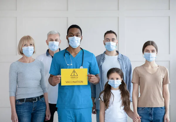 Portret różnych osób w maskach twarzy stojących za czarnym lekarzem ze znakiem szczepienia Covid-19 w klinice — Zdjęcie stockowe