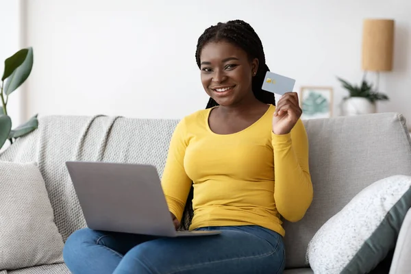 Pago fácil. Sonriente dama negra con computadora portátil y tarjeta de crédito en casa —  Fotos de Stock