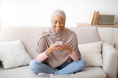 Hoş, tesettürlü siyahi kadın akıllı telefonuyla mesajlaşırken evdeki rahat koltukta oturuyor.