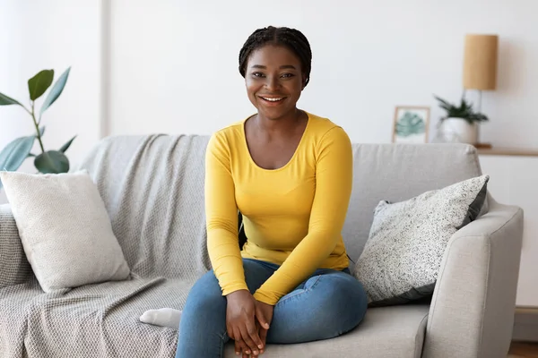 Mulher Millennial preta bonita sentada no sofá em casa, sorrindo para a câmera — Fotografia de Stock