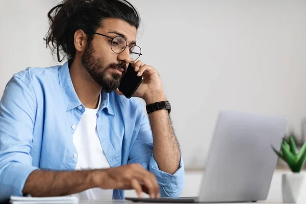 Beschäftigter junger arabischer Geschäftsmann, der mit dem Handy spricht und am Laptop arbeitet — Stockfoto